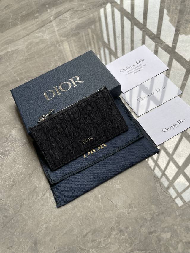 这款拉链卡夹是一款高雅经典的单品。采用米色和黑色Oblique印花面料精心制作，饰以黑色粒面牛皮革和Dior标志。顶部设有拉链隔层，背面设有四个卡槽。精巧实用，
