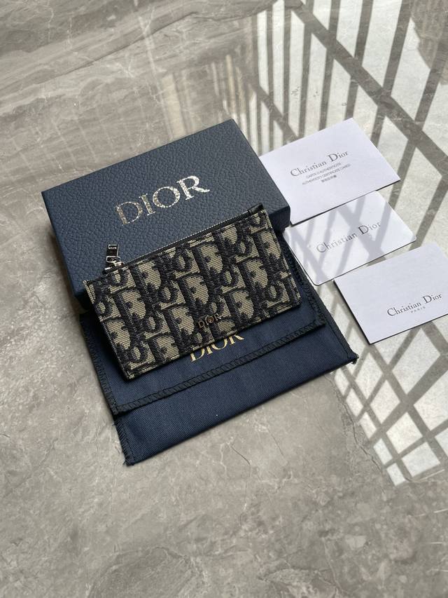这款拉链卡夹是一款高雅经典的单品。采用米色和黑色Oblique印花面料精心制作，饰以黑色粒面牛皮革和Dior标志。顶部设有拉链隔层，背面设有四个卡槽。精巧实用，