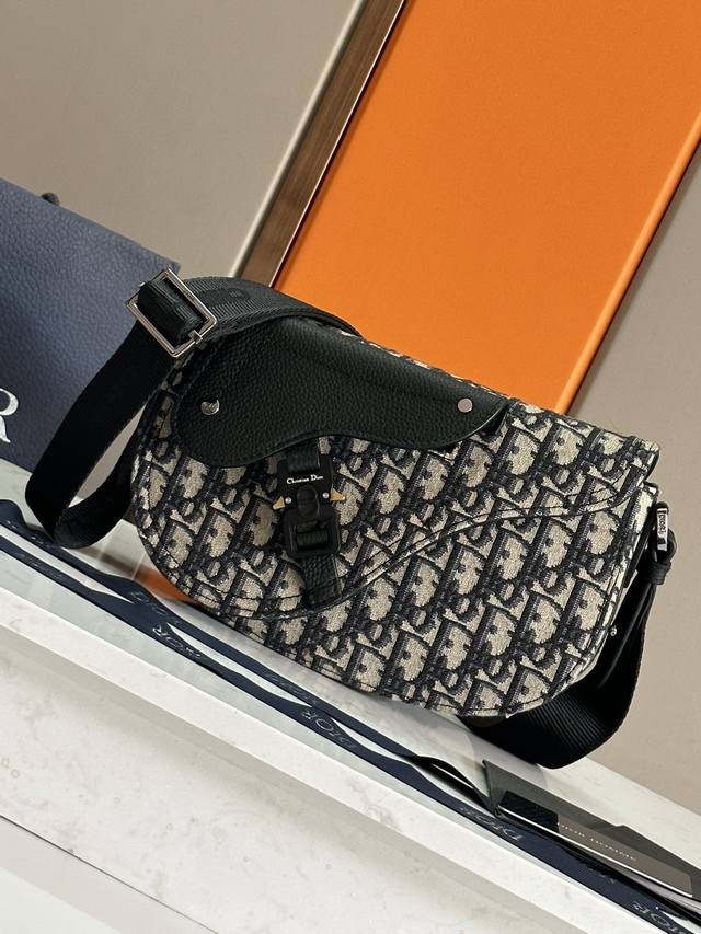 这款马鞍信使包是二零二四秋季男装系列新品，精巧时尚。采用米色和黑色 Oblique 印花面料精心制作，搭配黑色粒面牛皮革马鞍翻盖，点缀以铝扣提升格调。宽敞的内部