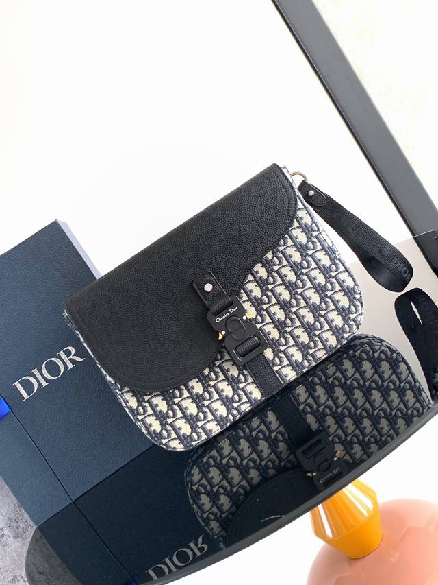 这款 A5 马鞍手拿包以时尚优雅的设计提升经典马鞍系列的格调。采用米色和黑色 Oblique 印花面料精心制作，搭配黑色粒面牛皮革马鞍翻盖，点缀以 Christ