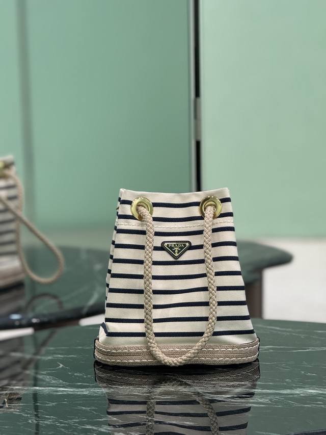 新款水桶包 这款织物水桶包饰以航海风条纹图案，为造型增添色彩。底部的设计灵感源自草编鞋，绳索则是象征着航海世界的另一细节元素，共同营造出浓郁的夏日风情。顶级五金