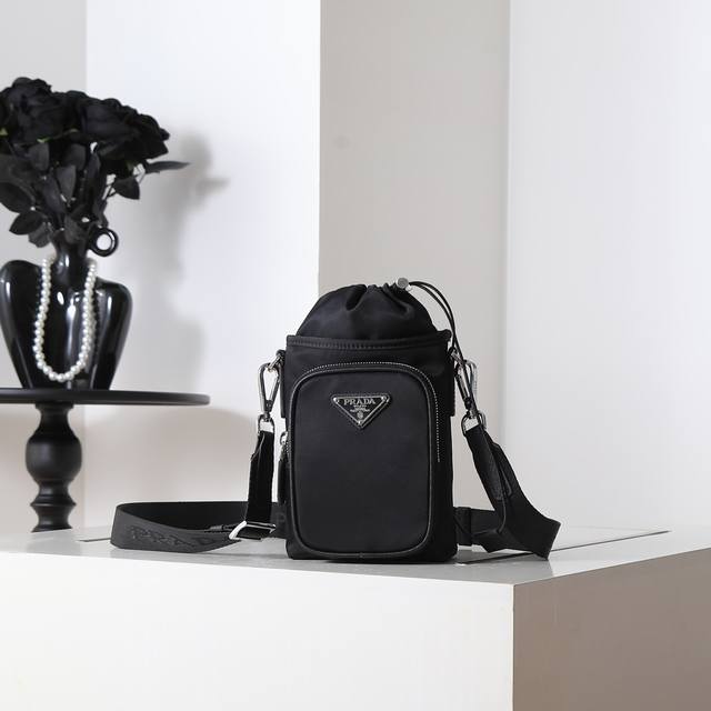 新款手机壳包2Zh155 这款手机包采用创新性Re-Nylon再生尼龙打造，展现充满现代感的都市风范。可调式尼龙织带肩带和束绳开合呈现别致特色。正面标志和前幅袋