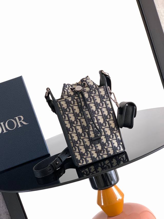 背景原厂 。这款水桶包是二零二五冬季男装系列新品，彰显时尚风范。挺括设计，采用米色和黑色 Oblique 印花面料精心制作，饰以黑色粒面牛皮革细节提升格调，正面